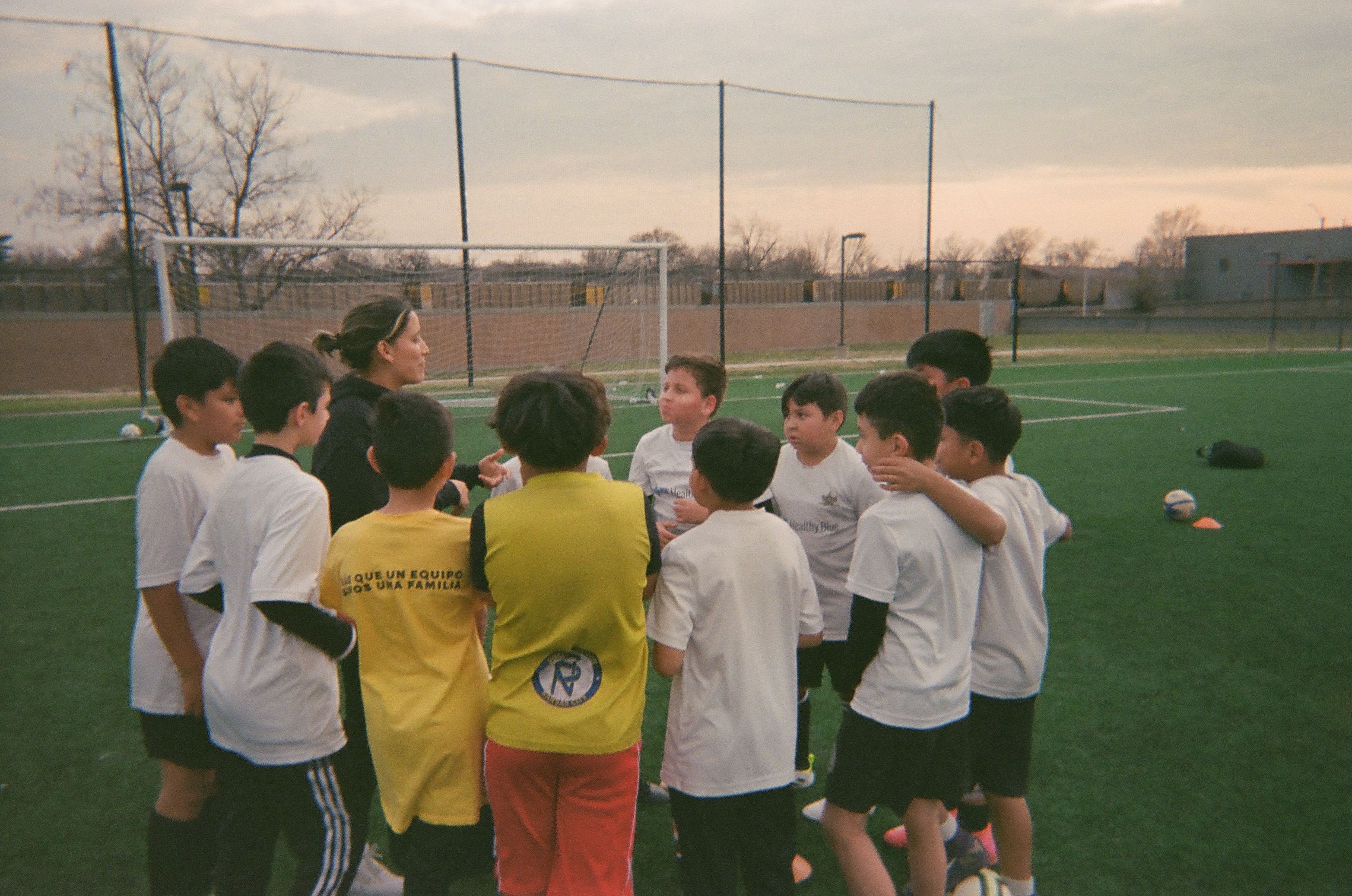 Group of children and a coach gathered in a huddle on a soccer pitch.