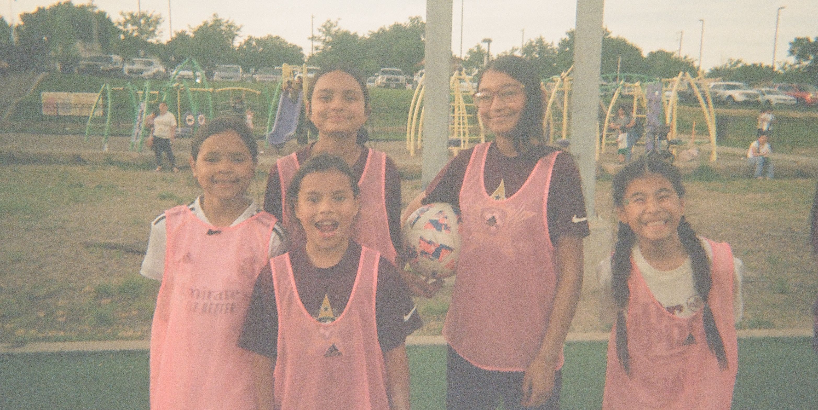 Five girls wearing training bibs smile together while holding a soccer ball at a community soccer field.