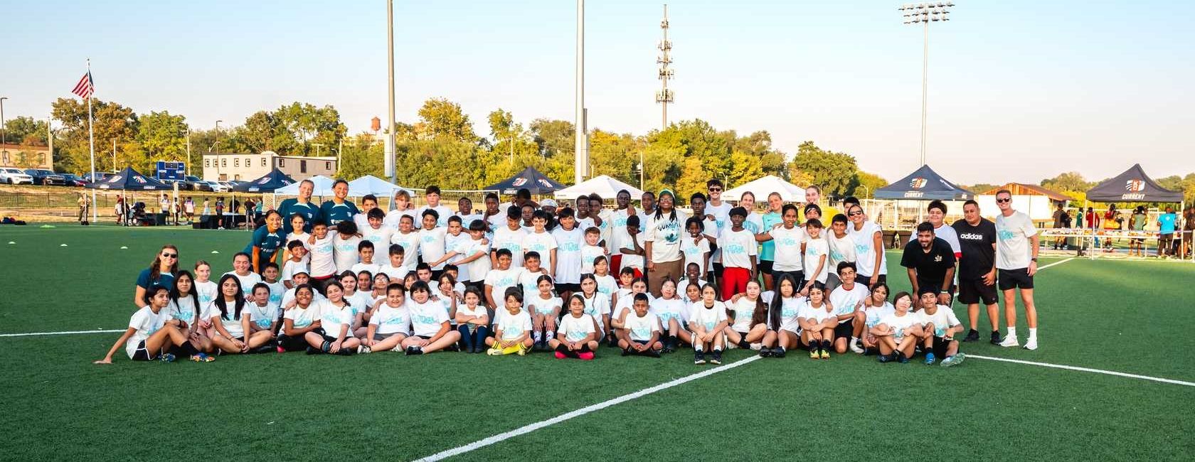 Group of children, coaches, and volunteers gathered for a photo on a soccer field during a Kansas City Current community soccer event.