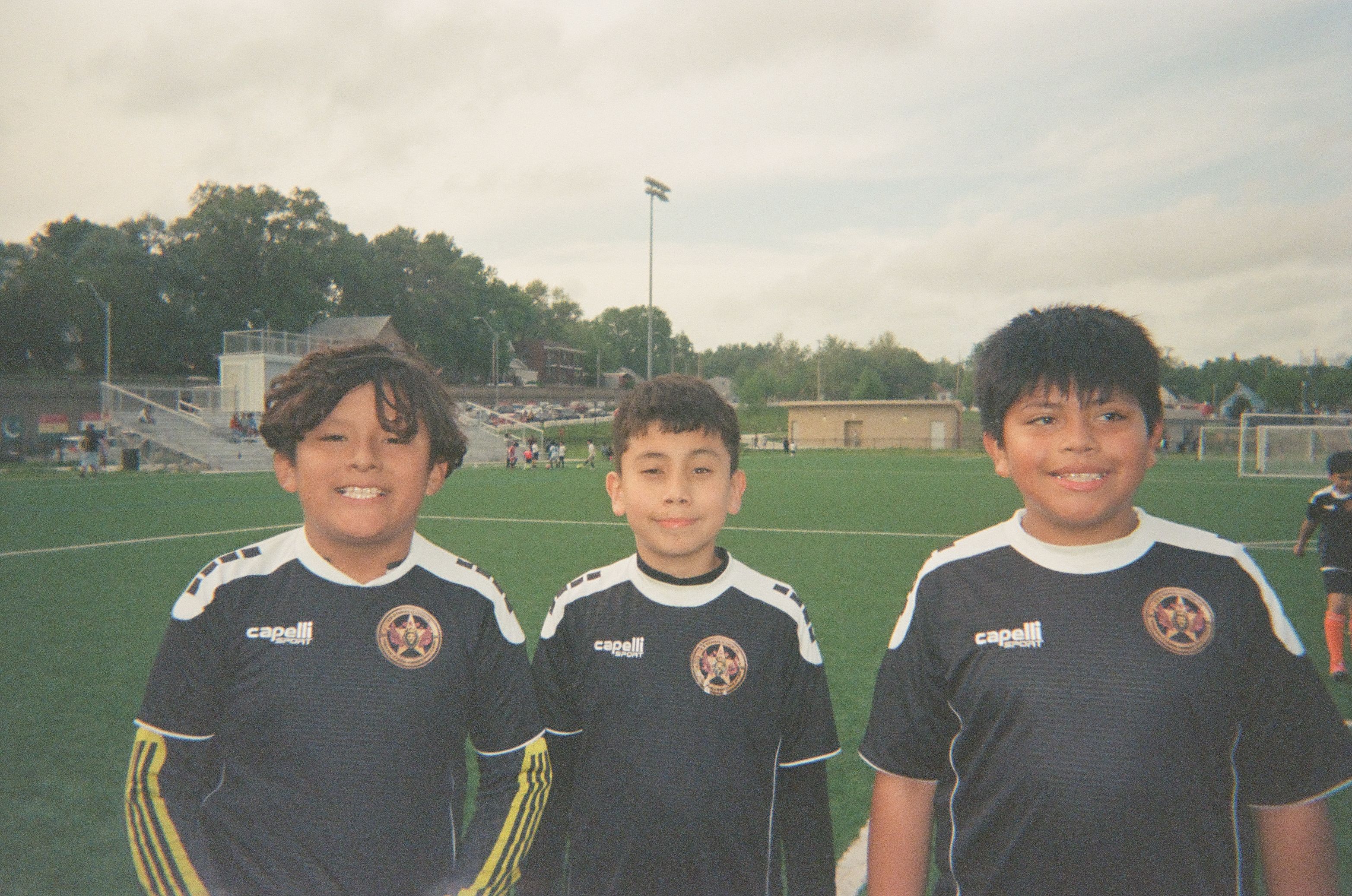 Three children wearing matching soccer jerseys stand together on an outdoor soccer field.