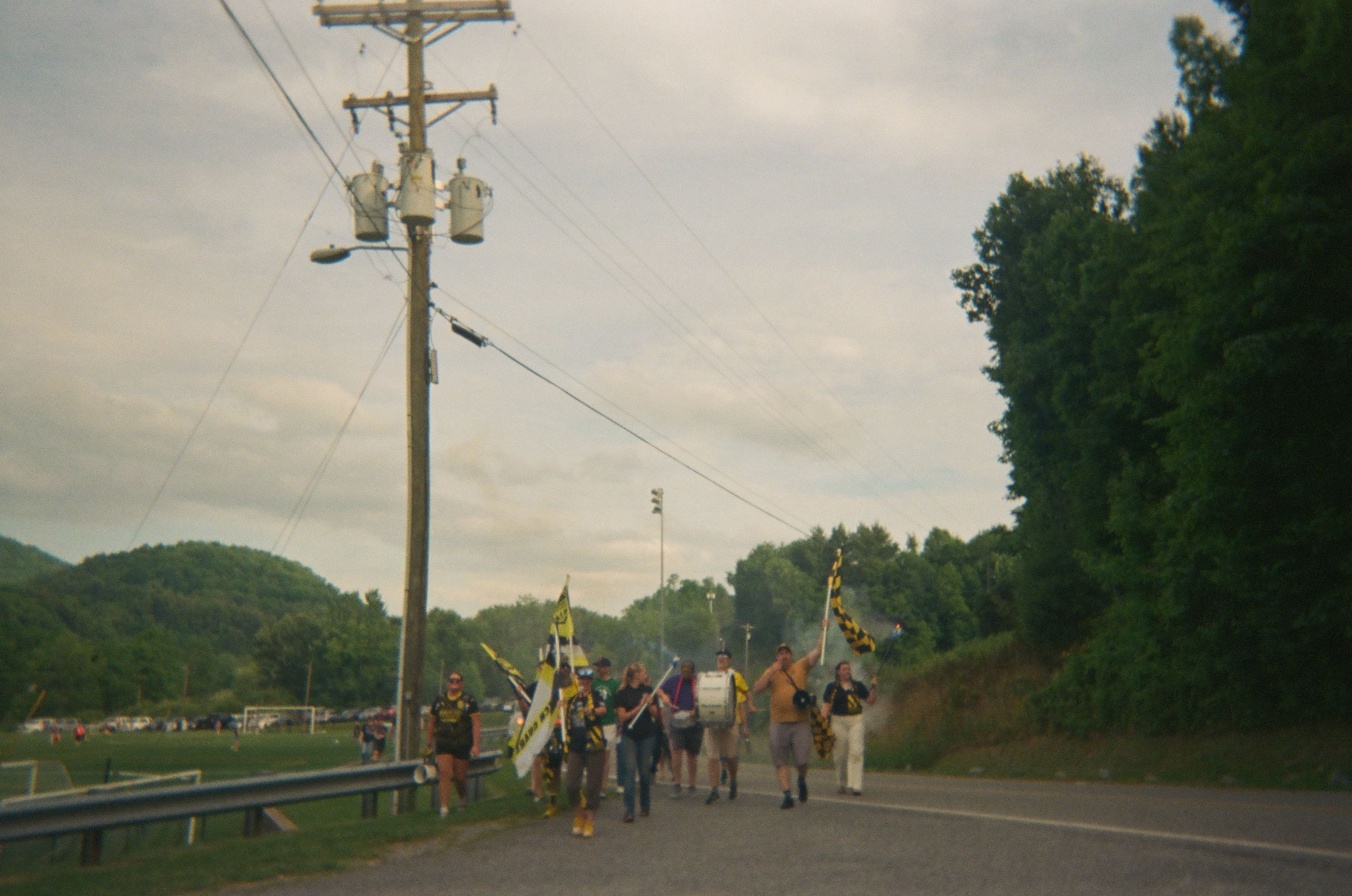 Soccer supporters walk along a road beside a field, carrying yellow and black flags and a drum under a cloudy sky.