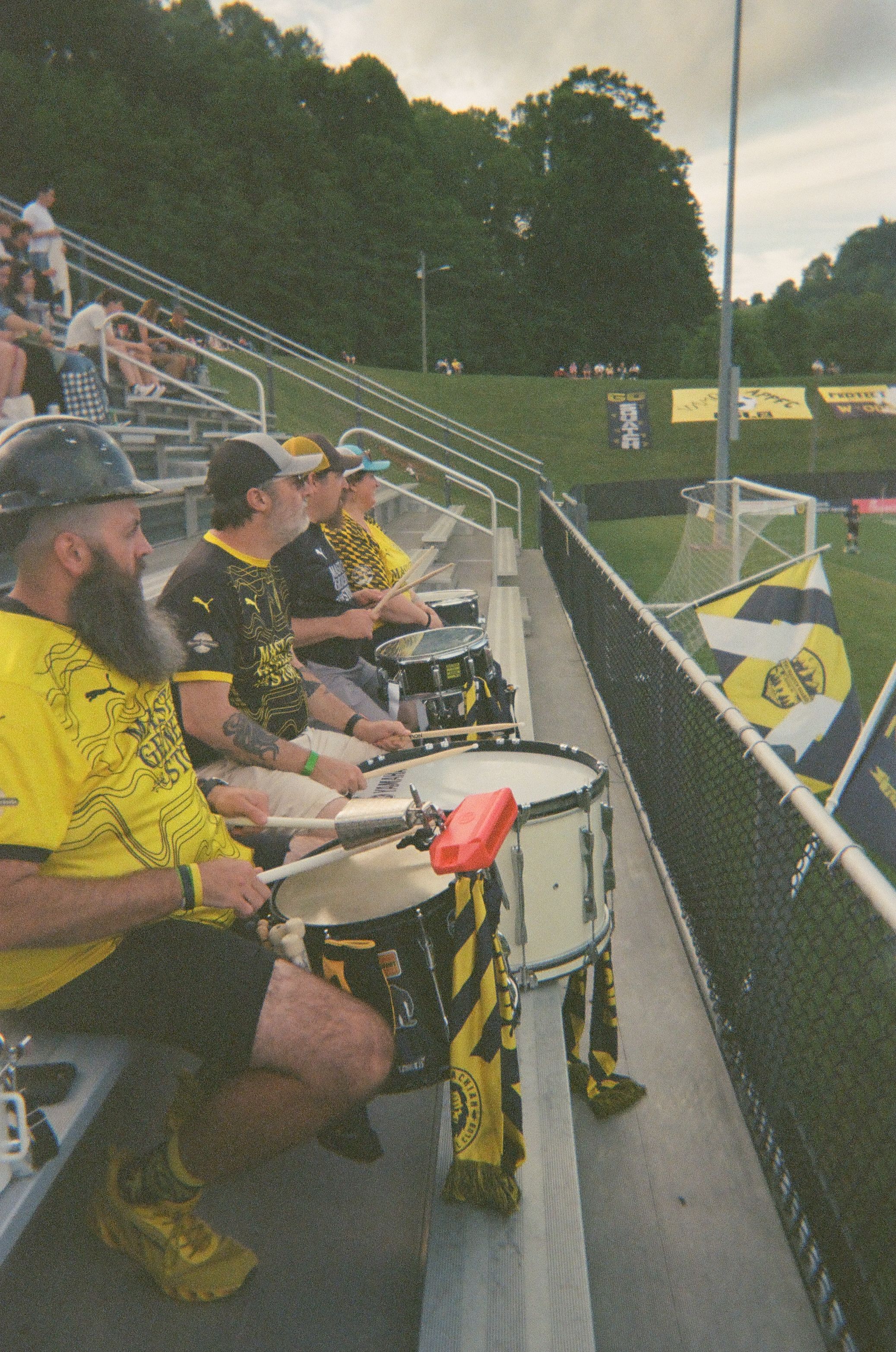 Supporters in yellow and black play drums in the bleachers beside a soccer field.