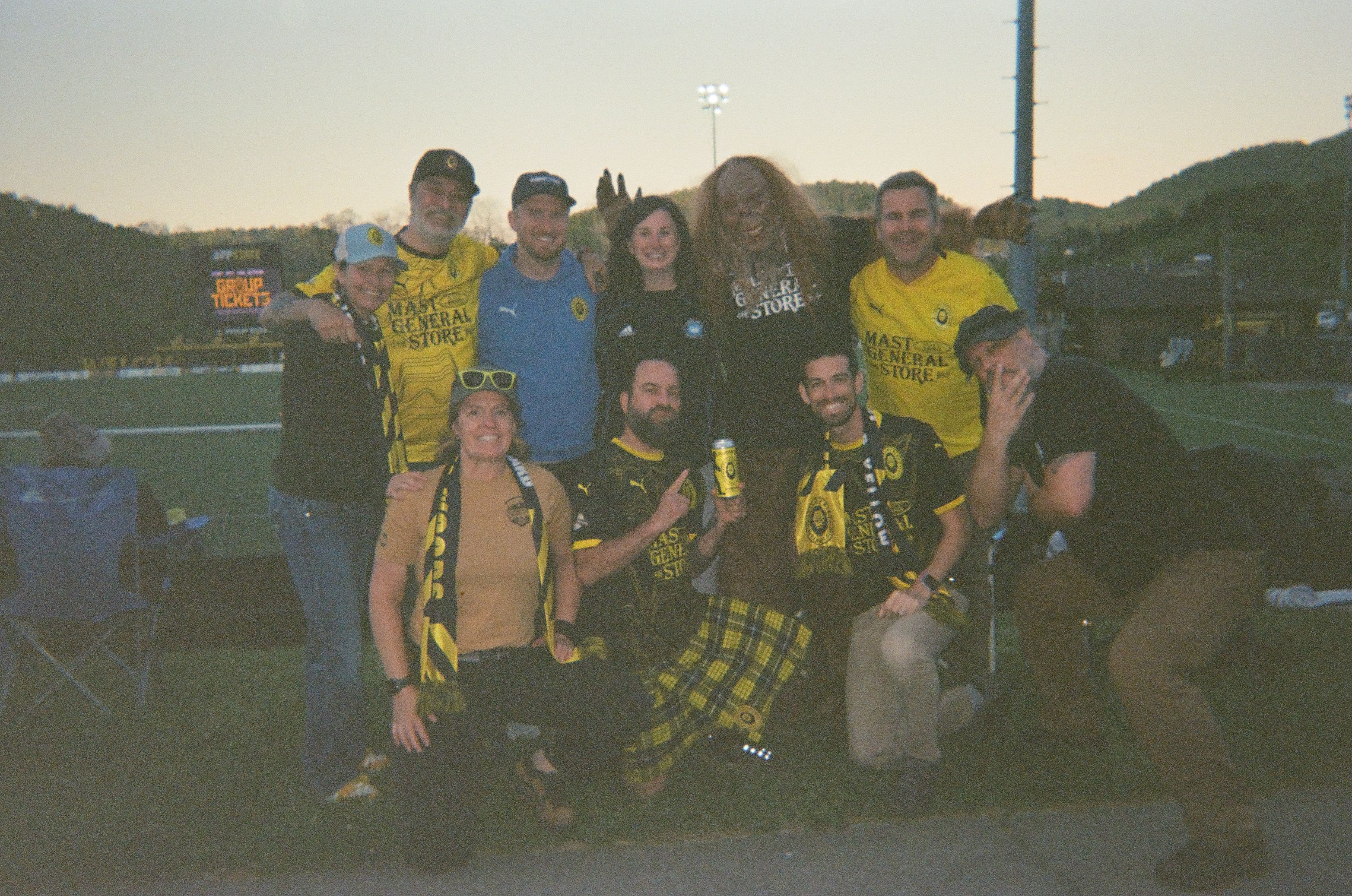 A group of nine soccer fans pose together beside a field at dusk, many wearing yellow and black team jerseys and scarves.