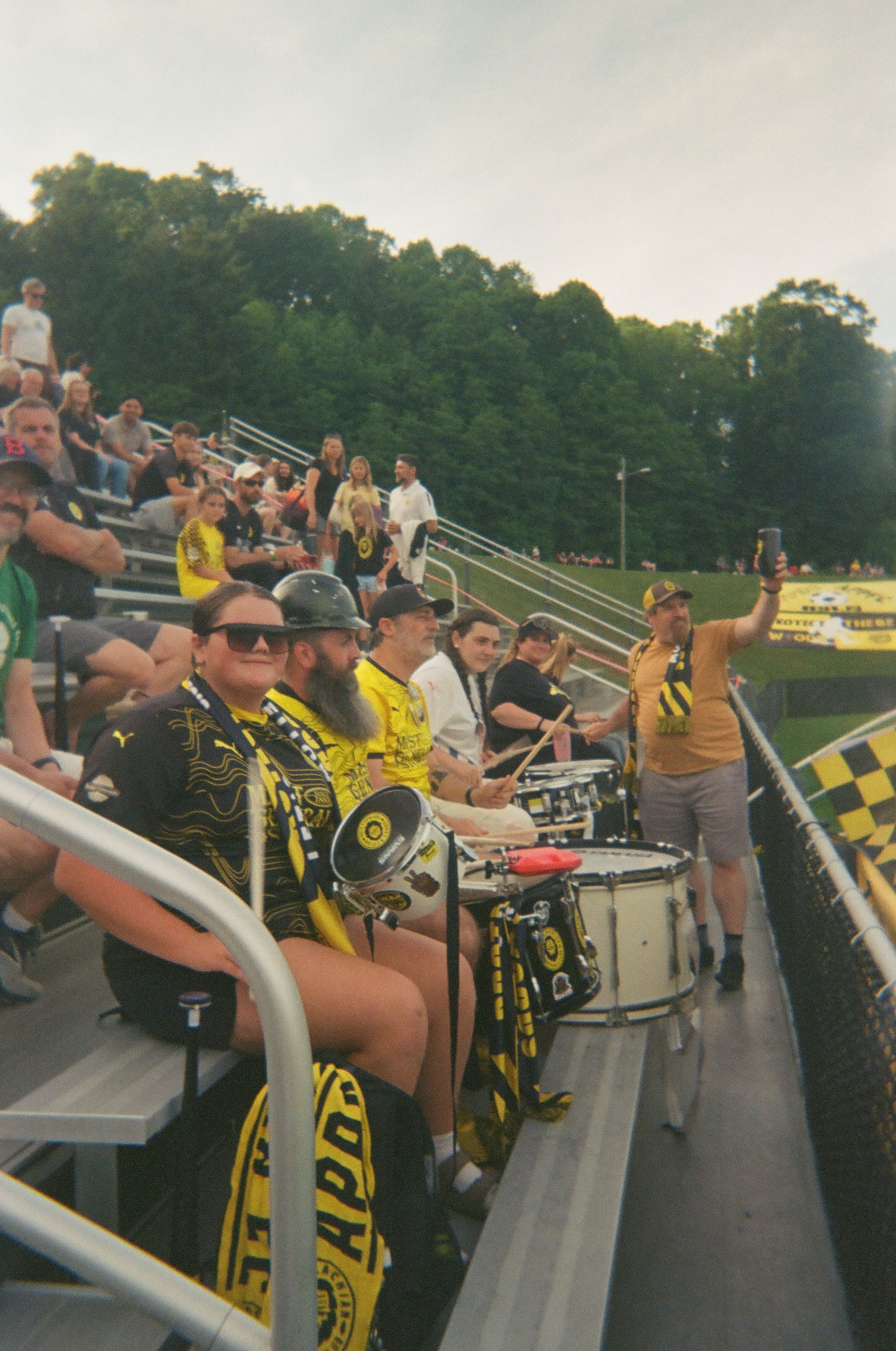 Soccer fans in yellow and black sit in the bleachers with drums and scarves.