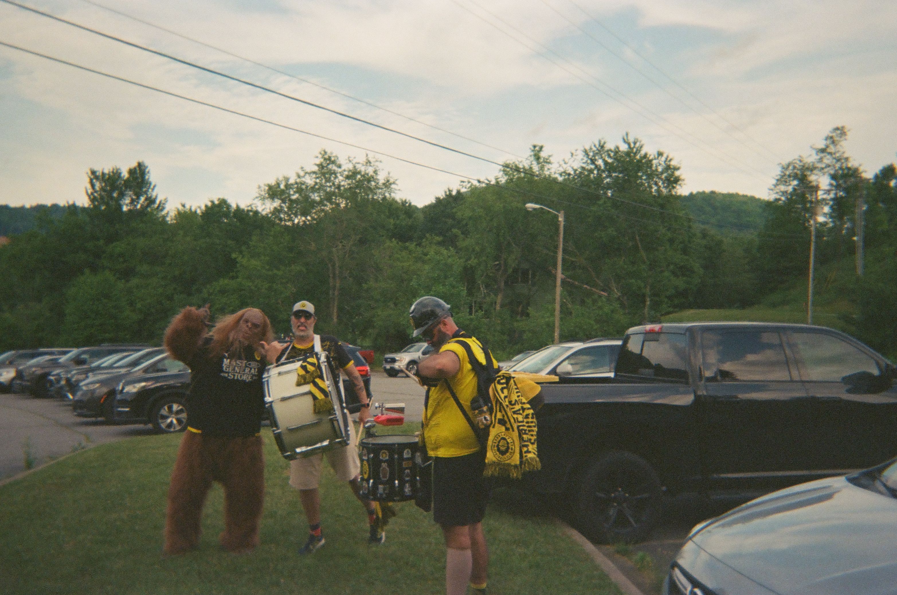 A soccer mascott walks with two drummers through a parking lot