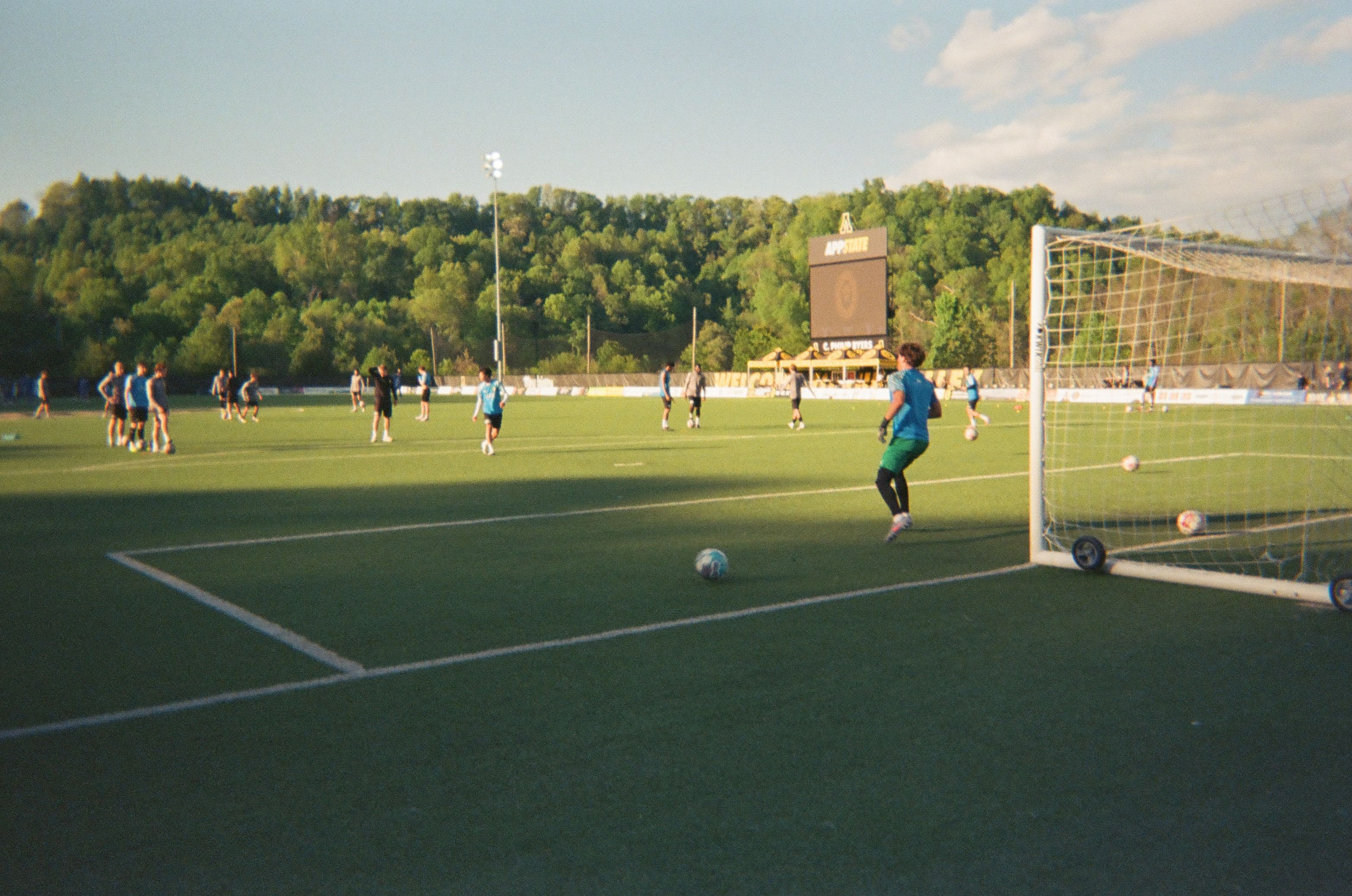 Soccer players warm up on a field as a goalkeeper stands beside the goal.