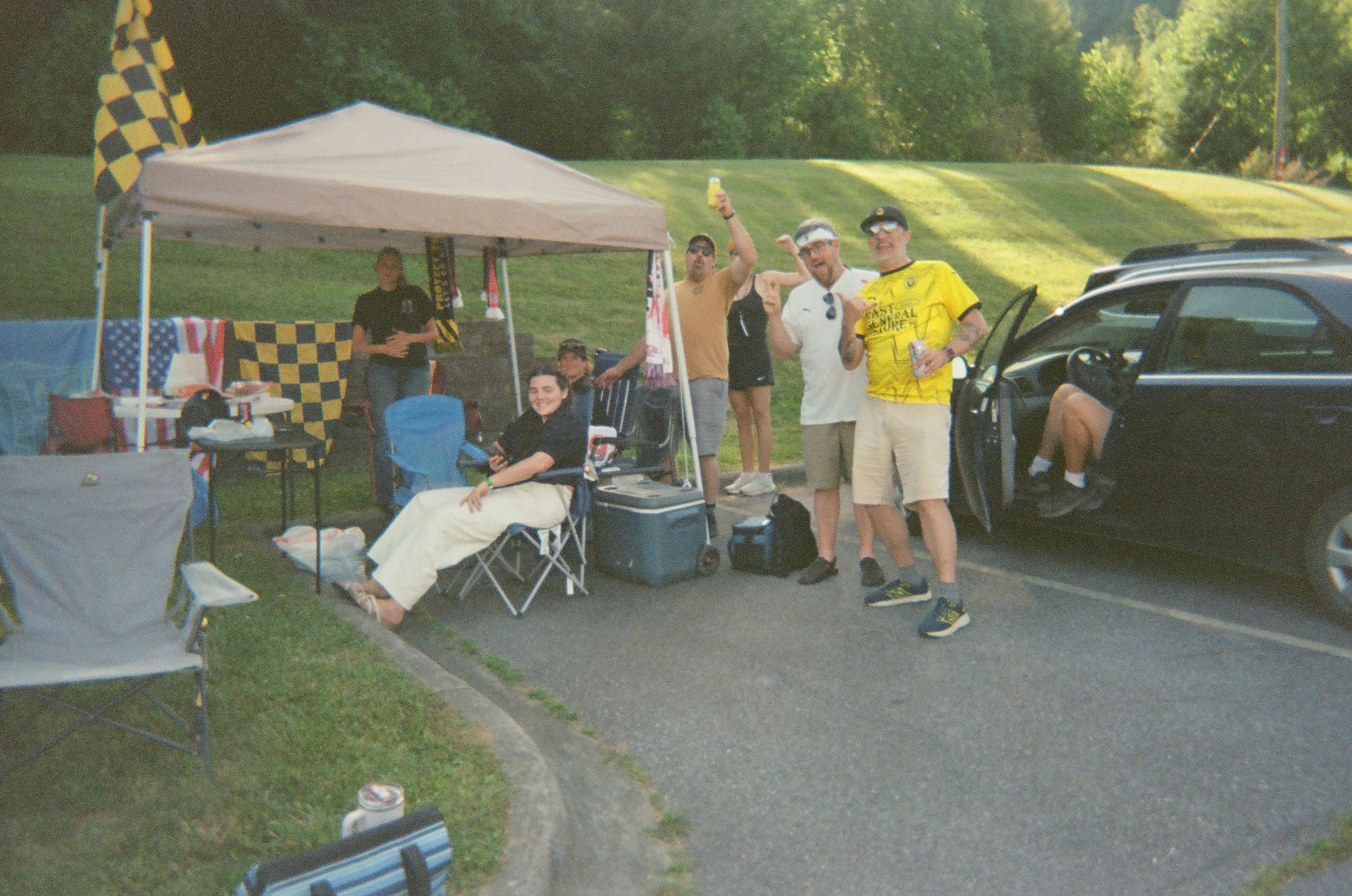 Soccer supporters gather beside a canopy and car, waving to the camera with yellow and black flags behind them.