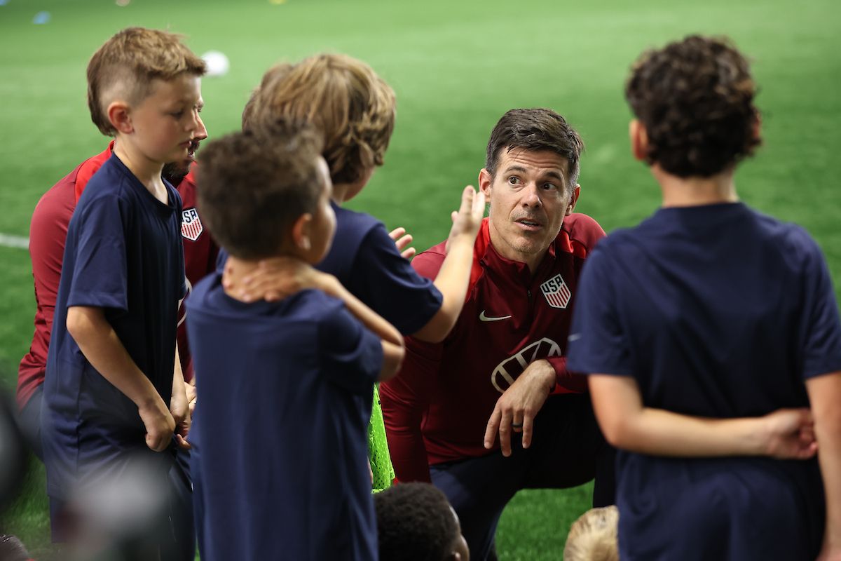 A coach kneels to speak with a group of young soccer players