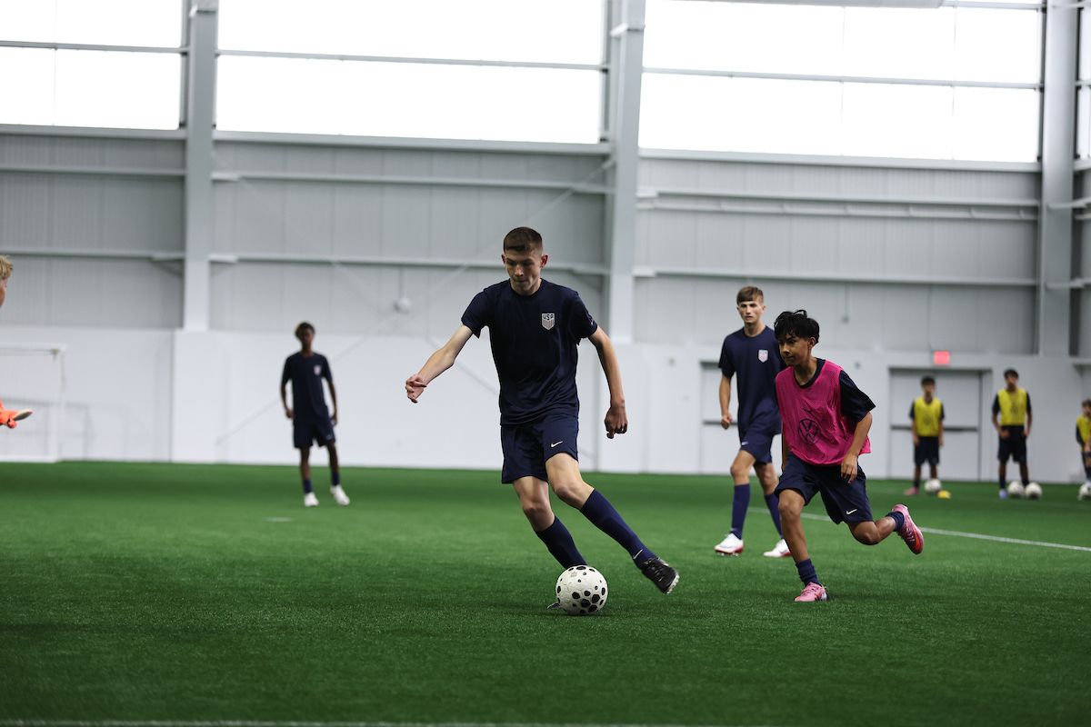 Young player in U.S. Soccer kit dribbles ball past a defender indoors.