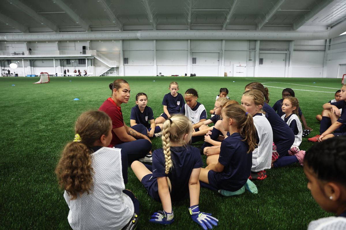 A coach sits with a group of young girls on an indoor soccer field.