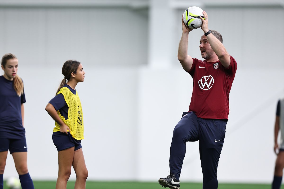 A coach demonstrates a skill with a ball overhead as a young player watches and learns.