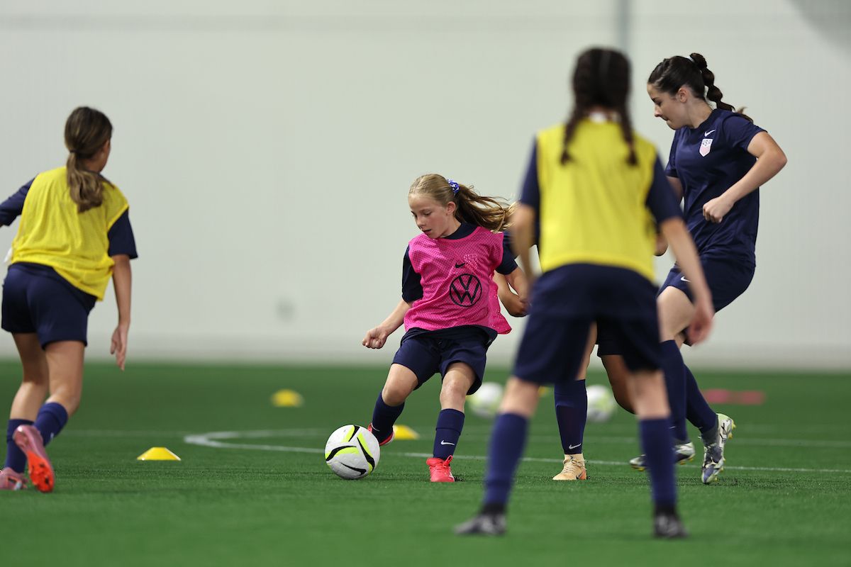 Young girl dribbles a soccer ball through defenders during indoor training.