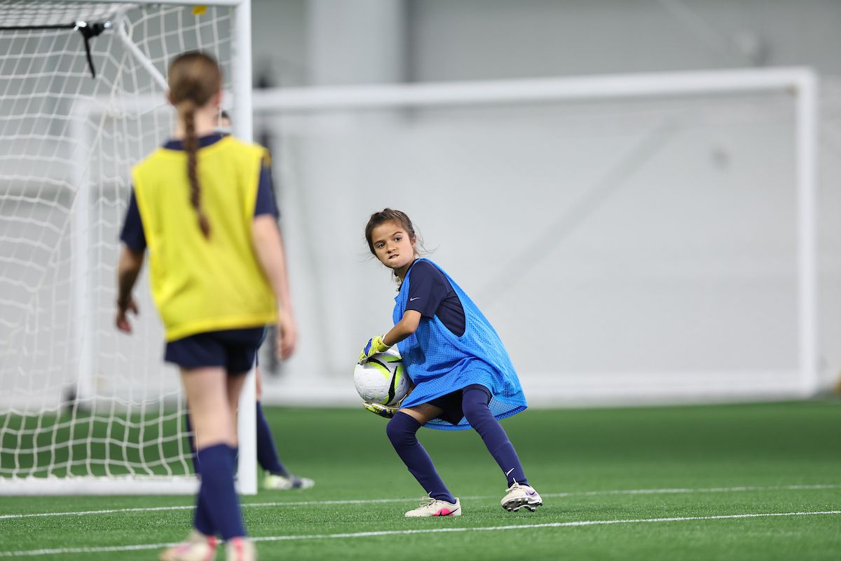 Young girl goalkeeper crouches to collect the ball with focused intensity.