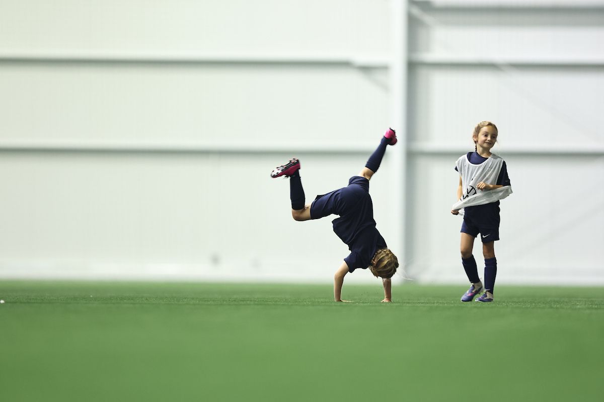 Young player doing a cartwheel on a soccer field while a teammate watches