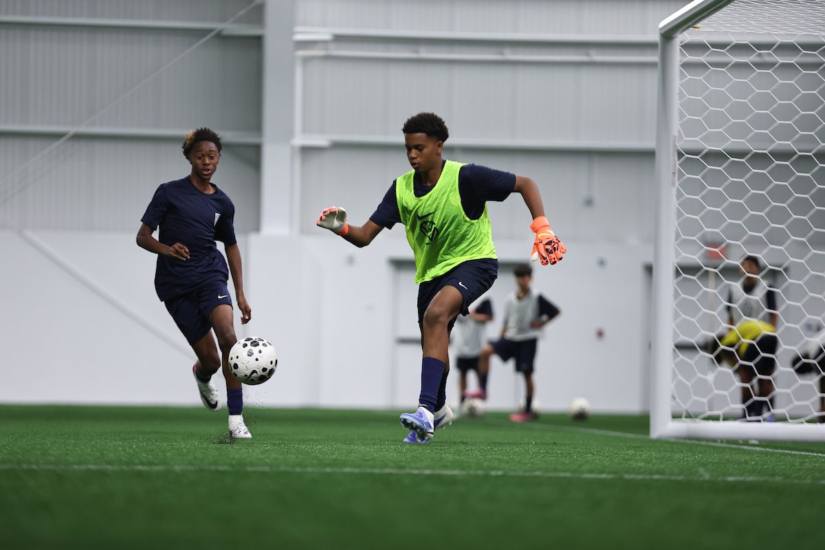 Young girl goalkeeper crouches to collect the ball with focused intensity.