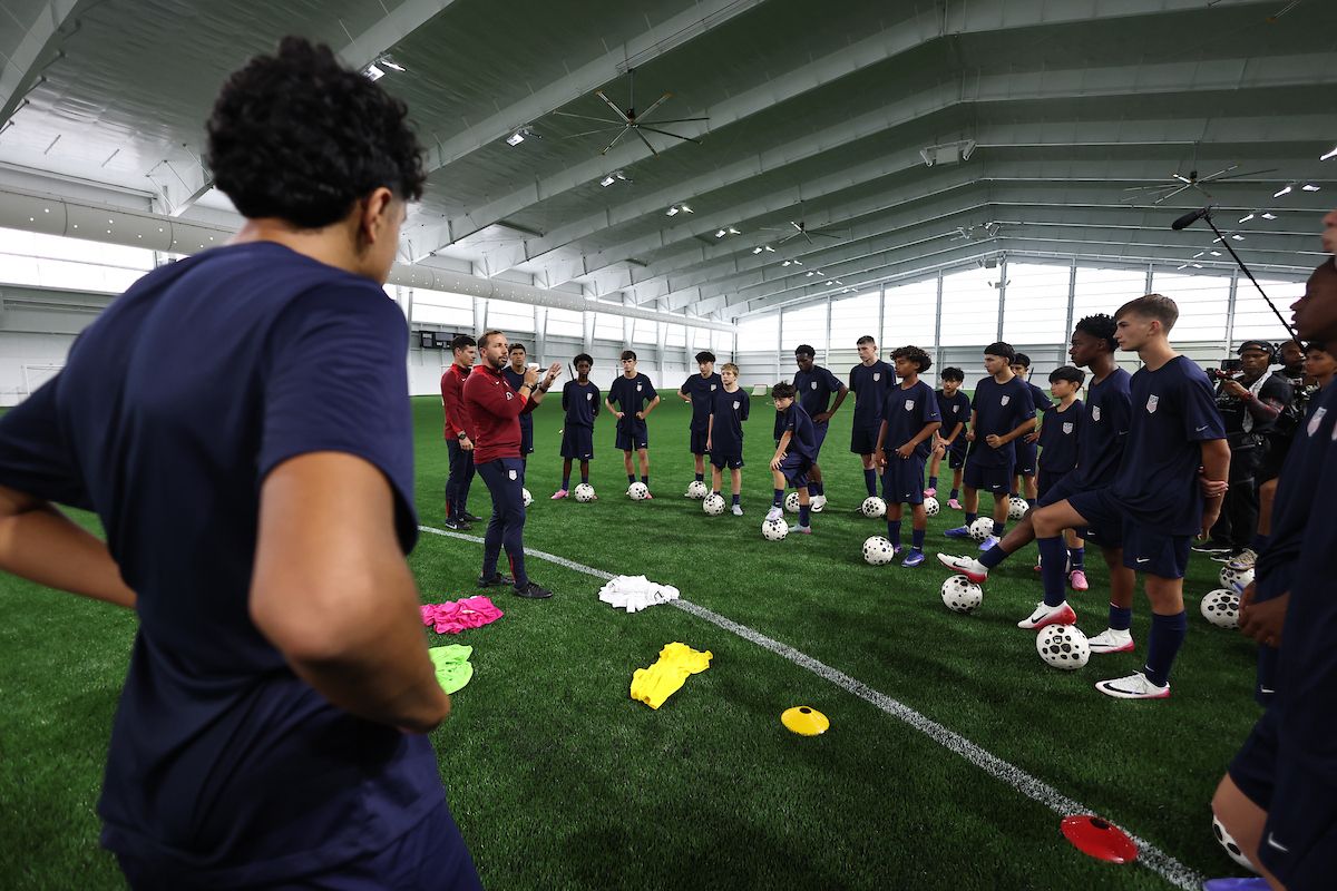 Coach addresses a group of youth soccer players gathered on an indoor field.