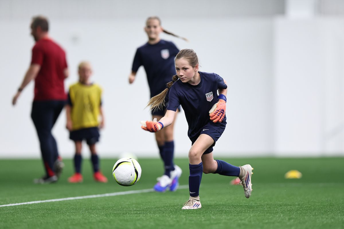 Young goalkeeper in U.S. Soccer kit lunges toward a soccer ball during training.
